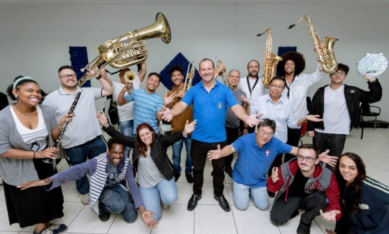 A imagem mostra um grupo grande e animado de músicos, muitos segurando seus instrumentos, sorrindo para a câmera. Há uma mistura de instrumentos de sopro, incluindo trompetes, saxofones, e um tuba. Um homem de camisa azul está no centro da foto, com os braços abertos, como se estivesse conduzindo ou liderando o grupo. As pessoas ao redor dele parecem ser de diversas idades e etnias. A atmosfera é descontraída e festiva, sugerindo uma reunião de banda ou um ensaio. O fundo é simples, uma parede branca com um pouco de decoração, indicando um ambiente interno como um salão de ensaio ou uma sala de aula. A iluminação é uniforme, sem sombras fortes, contribuindo para a clareza da imagem.