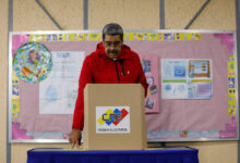 Um homem com um casaco vermelho está em frente a uma urna eleitoral, prestes a votar. Ao fundo, há um painel rosa com informações e gráficos, além de um objeto redondo colorido. A mesa onde ele está se apoiando é coberta por um tecido azul.