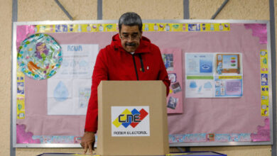 Um homem com um casaco vermelho está em frente a uma urna eleitoral, prestes a votar. Ao fundo, há um painel rosa com informações e gráficos, além de um objeto redondo colorido. A mesa onde ele está se apoiando é coberta por um tecido azul.