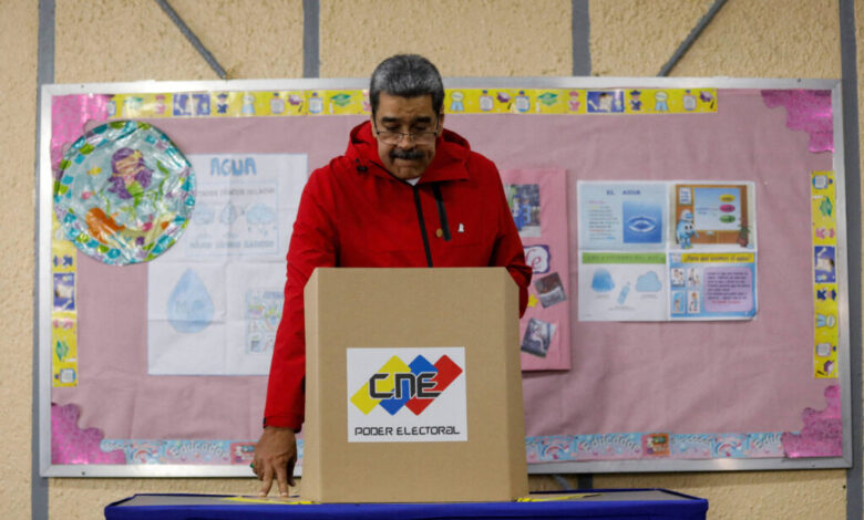 Um homem com um casaco vermelho está em frente a uma urna eleitoral, prestes a votar. Ao fundo, há um painel rosa com informações e gráficos, além de um objeto redondo colorido. A mesa onde ele está se apoiando é coberta por um tecido azul.