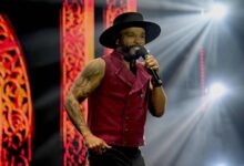 Um artista está se apresentando em um palco iluminado. Ele usa um colete vermelho e um chapéu preto, segurando um microfone. O fundo apresenta luzes brilhantes e elementos decorativos em tons de vermelho e dourado.