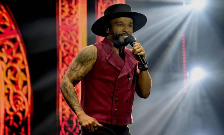 Um artista está se apresentando em um palco iluminado. Ele usa um colete vermelho e um chapéu preto, segurando um microfone. O fundo apresenta luzes brilhantes e elementos decorativos em tons de vermelho e dourado.