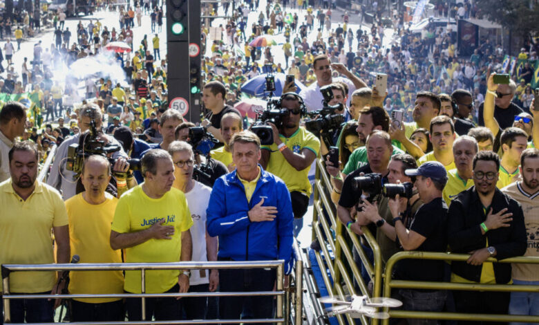 A imagem mostra uma grande multidão em uma manifestação ao ar livre. No primeiro plano, há um grupo de homens, alguns usando camisetas amarelas e outros com uma camiseta azul. Um dos homens, que está no centro, parece estar prestando atenção a algo à sua frente. Ao fundo, é possível ver uma grande quantidade de pessoas, muitas delas segurando bandeiras do Brasil. Há também várias câmeras e jornalistas registrando o evento.