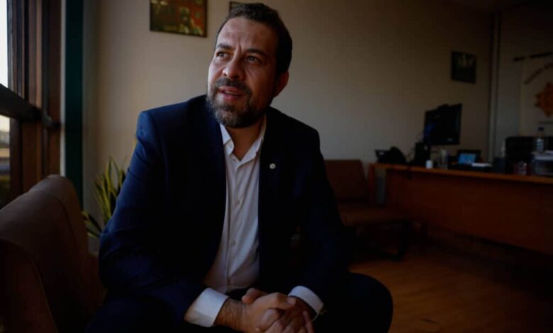 Um homem sentado em um ambiente de escritório, com uma expressão pensativa. Ele usa um terno escuro e uma camisa branca. Ao fundo, há uma mesa de escritório com um computador e algumas plantas. A luz natural entra pela janela, iluminando o espaço.