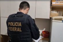 Um agente da Polícia Federal está de costas, usando um colete preto com a inscrição