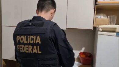 Um agente da Polícia Federal está de costas, usando um colete preto com a inscrição