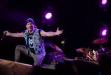 Um cantor se apresenta em um palco, usando um colete jeans e um gorro. Ele está em uma pose expressiva, com os braços abertos, enquanto um baterista é visível ao fundo. O cenário é iluminado por luzes roxas e há um fundo escuro.
