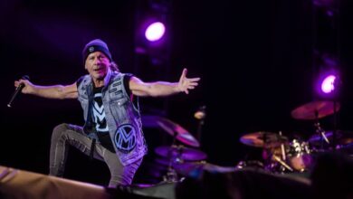 Um cantor se apresenta em um palco, usando um colete jeans e um gorro. Ele está em uma pose expressiva, com os braços abertos, enquanto um baterista é visível ao fundo. O cenário é iluminado por luzes roxas e há um fundo escuro.