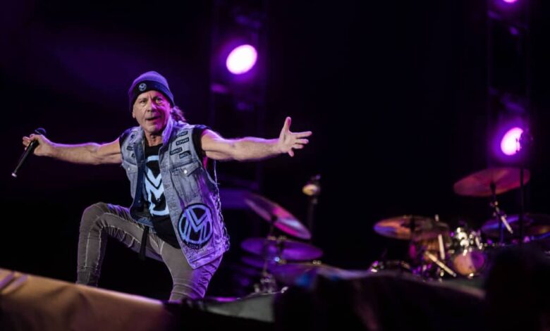 Um cantor se apresenta em um palco, usando um colete jeans e um gorro. Ele está em uma pose expressiva, com os braços abertos, enquanto um baterista é visível ao fundo. O cenário é iluminado por luzes roxas e há um fundo escuro.