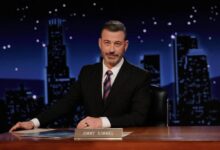 Um homem sentado em uma mesa de talk show, com um fundo noturno que mostra edifícios iluminados. Ele tem cabelo grisalho e usa um terno escuro com uma gravata listrada. Na mesa, há um nome em uma placa que diz