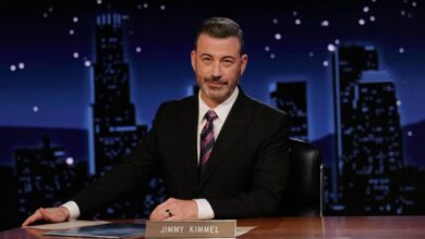 Um homem sentado em uma mesa de talk show, com um fundo noturno que mostra edifícios iluminados. Ele tem cabelo grisalho e usa um terno escuro com uma gravata listrada. Na mesa, há um nome em uma placa que diz