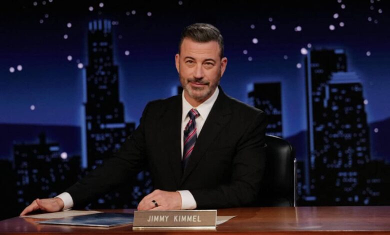 Um homem sentado em uma mesa de talk show, com um fundo noturno que mostra edifícios iluminados. Ele tem cabelo grisalho e usa um terno escuro com uma gravata listrada. Na mesa, há um nome em uma placa que diz