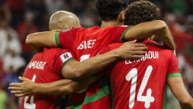 Três jogadores de futebol de Marrocos estão se abraçando em um momento de comemoração. Eles vestem uniformes vermelhos com detalhes verdes. O jogador à esquerda tem o número 4 nas costas, o do meio tem o número 7 e o da direita tem o número 14. O fundo mostra uma multidão, sugerindo um ambiente de estádio.