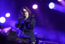 Uma artista feminina está se apresentando em um palco iluminado com luzes roxas e azuis. Ela está usando um traje escuro e segurando um microfone, enquanto faz gestos expressivos com as mãos. O fundo mostra uma configuração de palco com equipamentos e luzes.