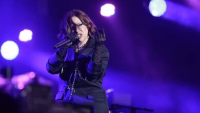 Uma artista feminina está se apresentando em um palco iluminado com luzes roxas e azuis. Ela está usando um traje escuro e segurando um microfone, enquanto faz gestos expressivos com as mãos. O fundo mostra uma configuração de palco com equipamentos e luzes.