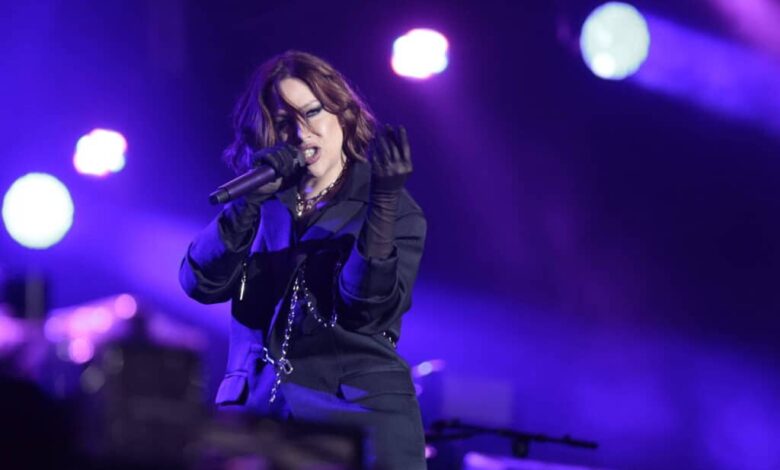 Uma artista feminina está se apresentando em um palco iluminado com luzes roxas e azuis. Ela está usando um traje escuro e segurando um microfone, enquanto faz gestos expressivos com as mãos. O fundo mostra uma configuração de palco com equipamentos e luzes.